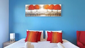 A hotel room with a blue wall, a painting of trees, two red pillows, a white bedspread, and a red chair. - Ocean Breeze 59 (Cape Town)