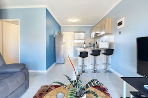 A modern living room with a grey sofa, a coffee table with decorative items, a kitchen area with a fridge, microwave, and bar stools, and a TV on the right.