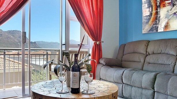 A living room with a round wooden coffee table, a sofa, and a large window with red curtains. - Ocean Breeze 59 (Cape Town)