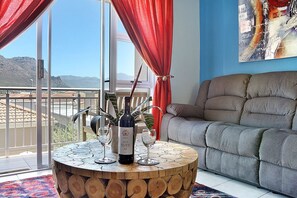 A living room with a round wooden coffee table, a sofa, and a large window with red curtains. - Ocean Breeze 59 (Cape Town)