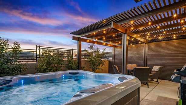 A hot tub under a pergola with string lights, surrounded by patio furniture and plants.