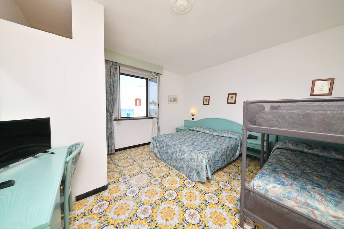 A room with a bunk bed, a desk with a television, a window with curtains, and patterned floor tiles.