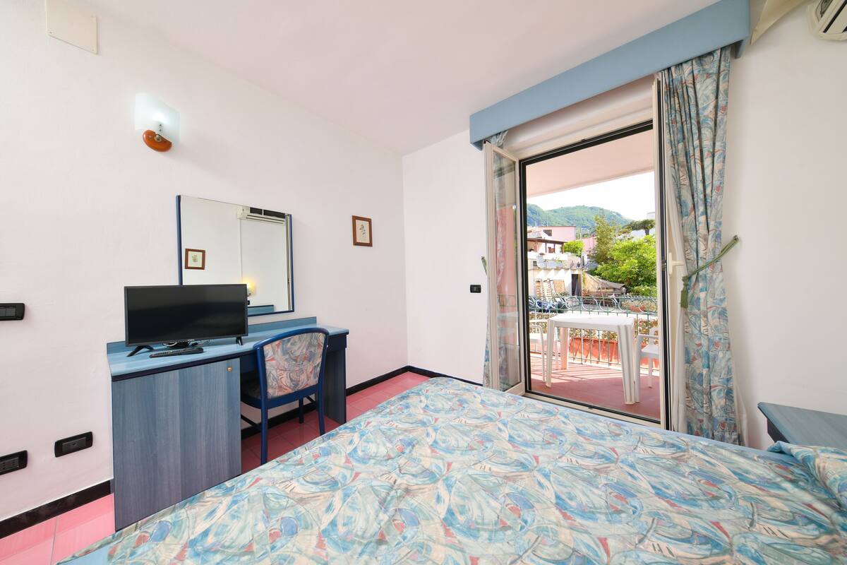 A hotel room with a bed, a desk with a computer, a chair, a window with a view of the outside, and a balcony.