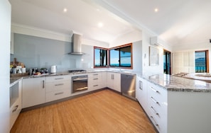 A modern kitchen with white cabinetry, a granite countertop, and a view of the ocean through large windows. - Coral Sea Views (Airlie Beach)