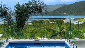 Piscina com guarda-corpo de vidro, cercada por vegetação exuberante e vista para um lago e colinas.