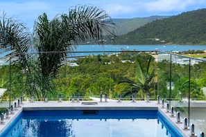 A swimming pool with a glass balustrade, surrounded by lush greenery and a view of a lake and hills. - Coral Sea Views (Airlie Beach)