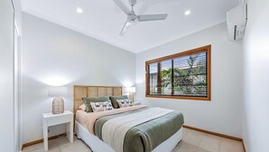Quarto com cama, mesa de cabeceira, luminária, janela com persianas, ventilador de teto e ar condicionado.