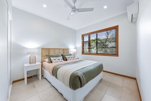 A bedroom with a bed, bedside table, lamp, window with blinds, ceiling fan, and air conditioning unit. - Coral Sea Views (Airlie Beach)