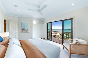 A bedroom with a bed, a ceiling fan, a large window with a view of the beach, and a balcony with outdoor furniture. - Coral Sea Views (Airlie Beach)