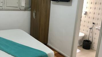 Quarto com cama, guarda-roupa, televisão e banheiro visível por uma porta aberta.