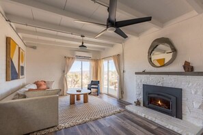 A spacious living room with a fireplace, a ceiling fan, and a large window with curtains. - Kinetic House by Homestead Modern (Yucca Valley)