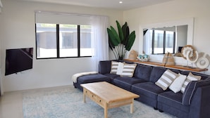 A living room with a dark blue sofa, a wooden coffee table, a flat-screen TV, and a large window with white curtains. - 301 THE LOOKOUT PENTHOUSE (South West Rocks)