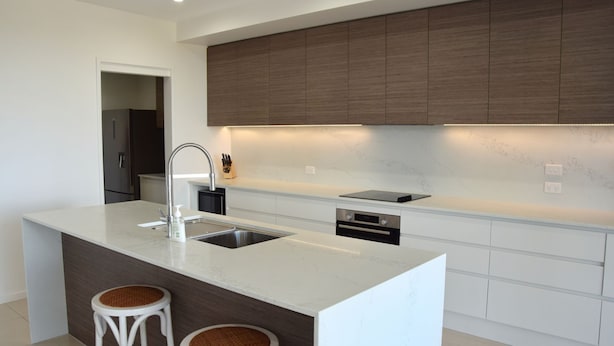 A modern kitchen with a central island, white cabinetry, and a dark wood accent wall. - 301 THE LOOKOUT PENTHOUSE (South West Rocks)