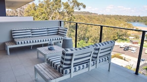 A balcony with a striped sofa set, a small table with a glass dome, and a view of a lake and surrounding greenery. - 301 THE LOOKOUT PENTHOUSE (South West Rocks)