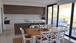 A modern kitchen with white cabinetry, a central island, and a dining area with white chairs and a wooden table. - 301 THE LOOKOUT PENTHOUSE (South West Rocks)
