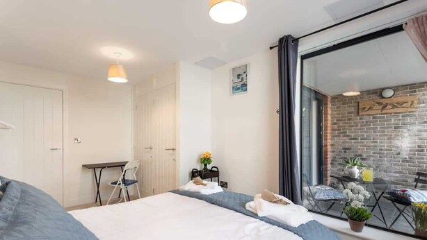 A hotel room with a large bed, a desk, a chair, and a balcony with a view of a brick wall and plants.