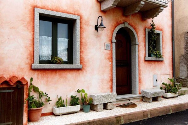 Un bâtiment à la façade rose, avec une porte en bois et une fenêtre ornée d’un rideau. Devant, on trouve des plantes en pot et un banc en pierre.