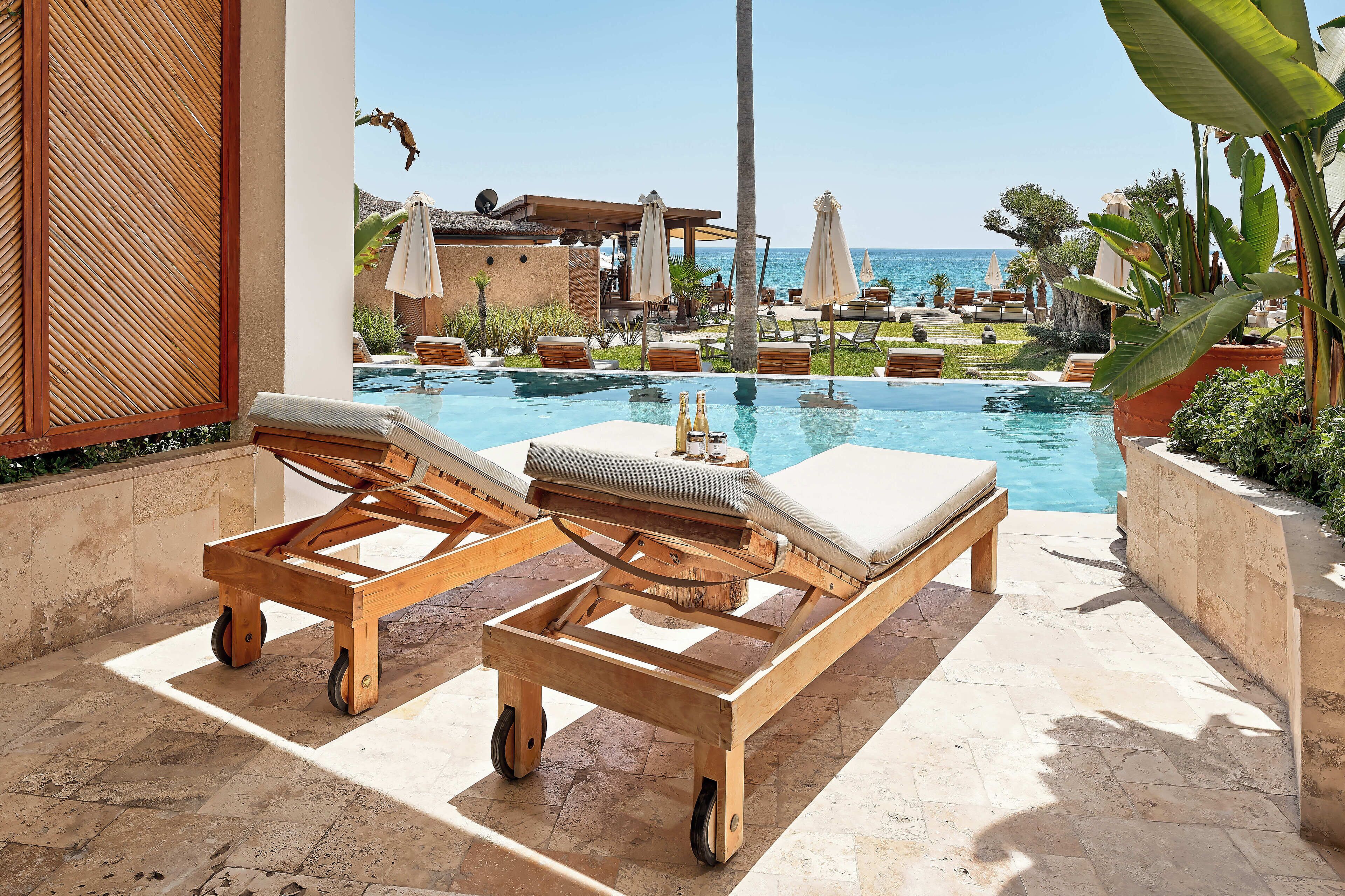 Zona junto a la piscina con sillones de descanso, un carrito de madera y vista al océano.