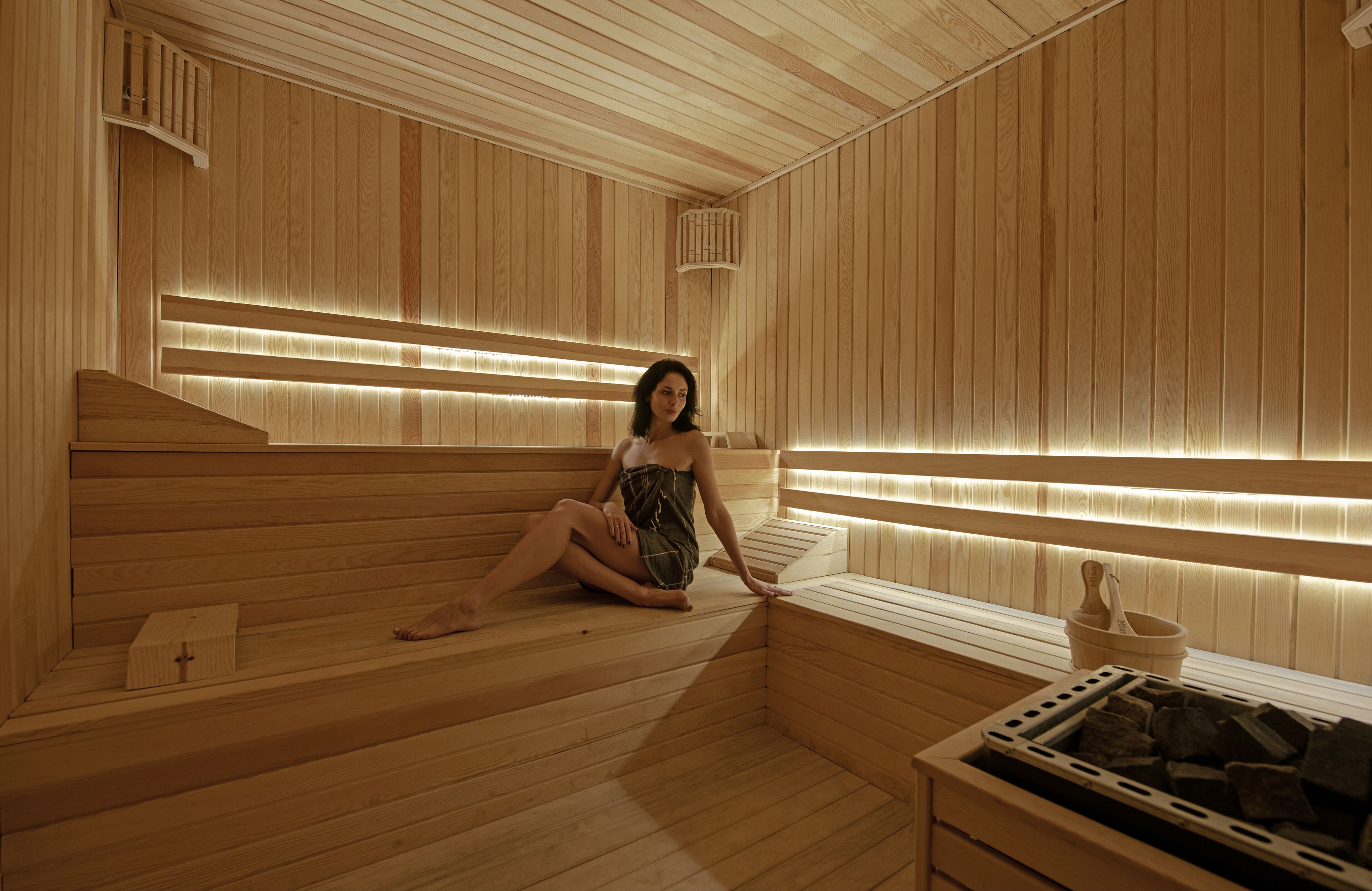 A sauna with wooden walls and benches, a small table with a bucket and stones, and a window with blinds.