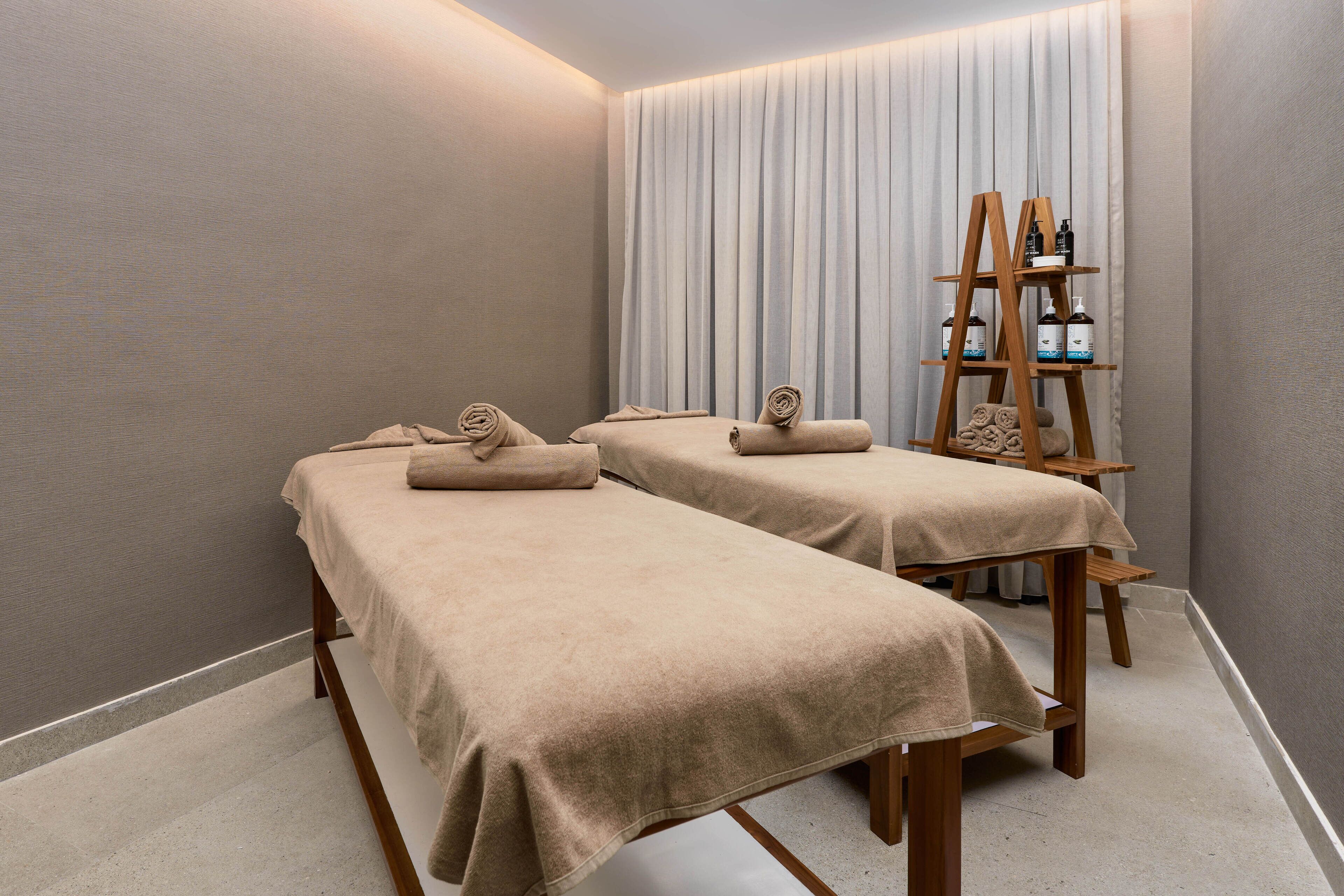 A spa room with two massage tables, a wooden shelf with bottles, and a large window with curtains.