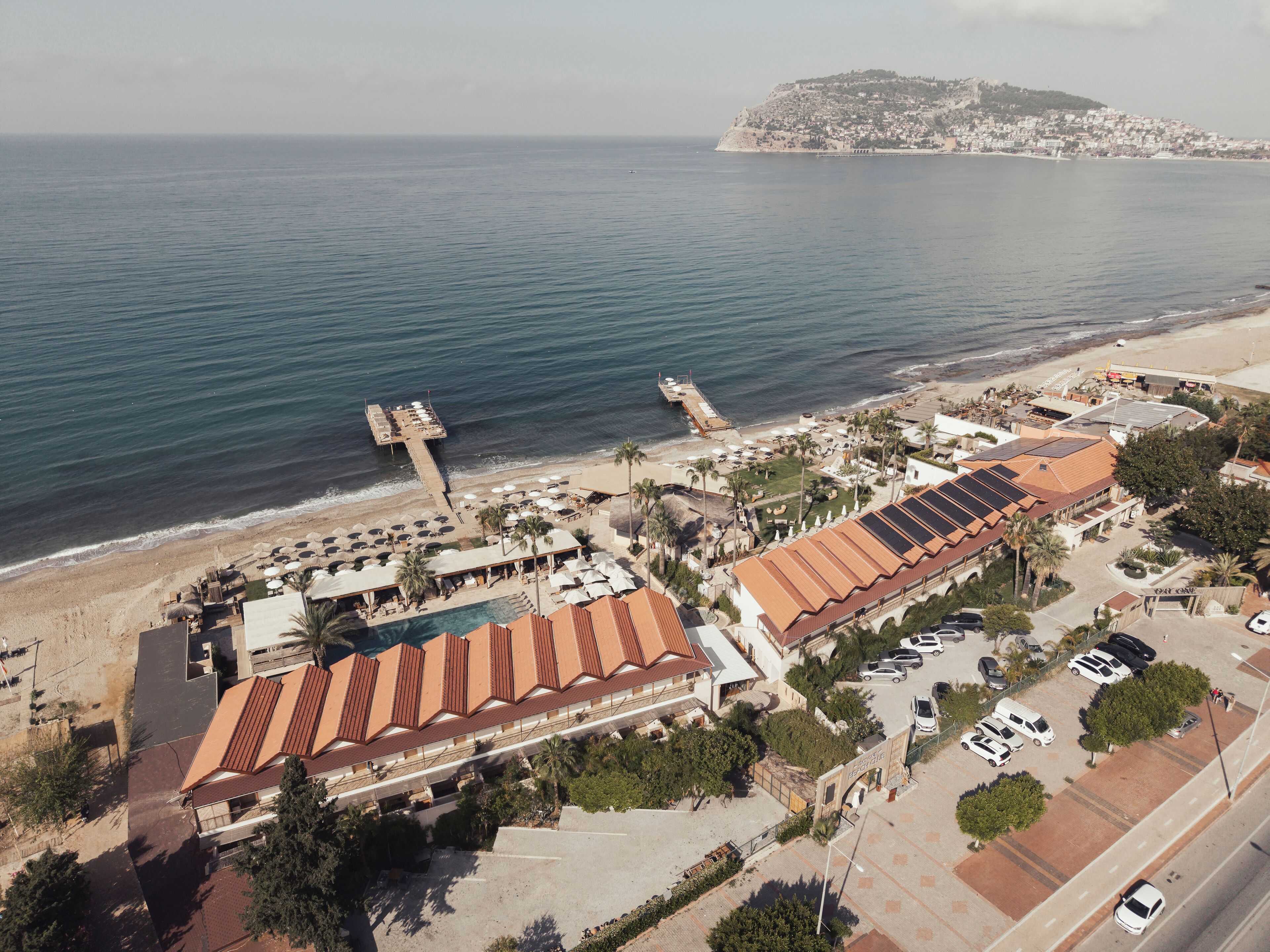 Vue aérienne d’un hôtel situé sur la côte, avec une plage, une jetée et une montagne en arrière-plan.