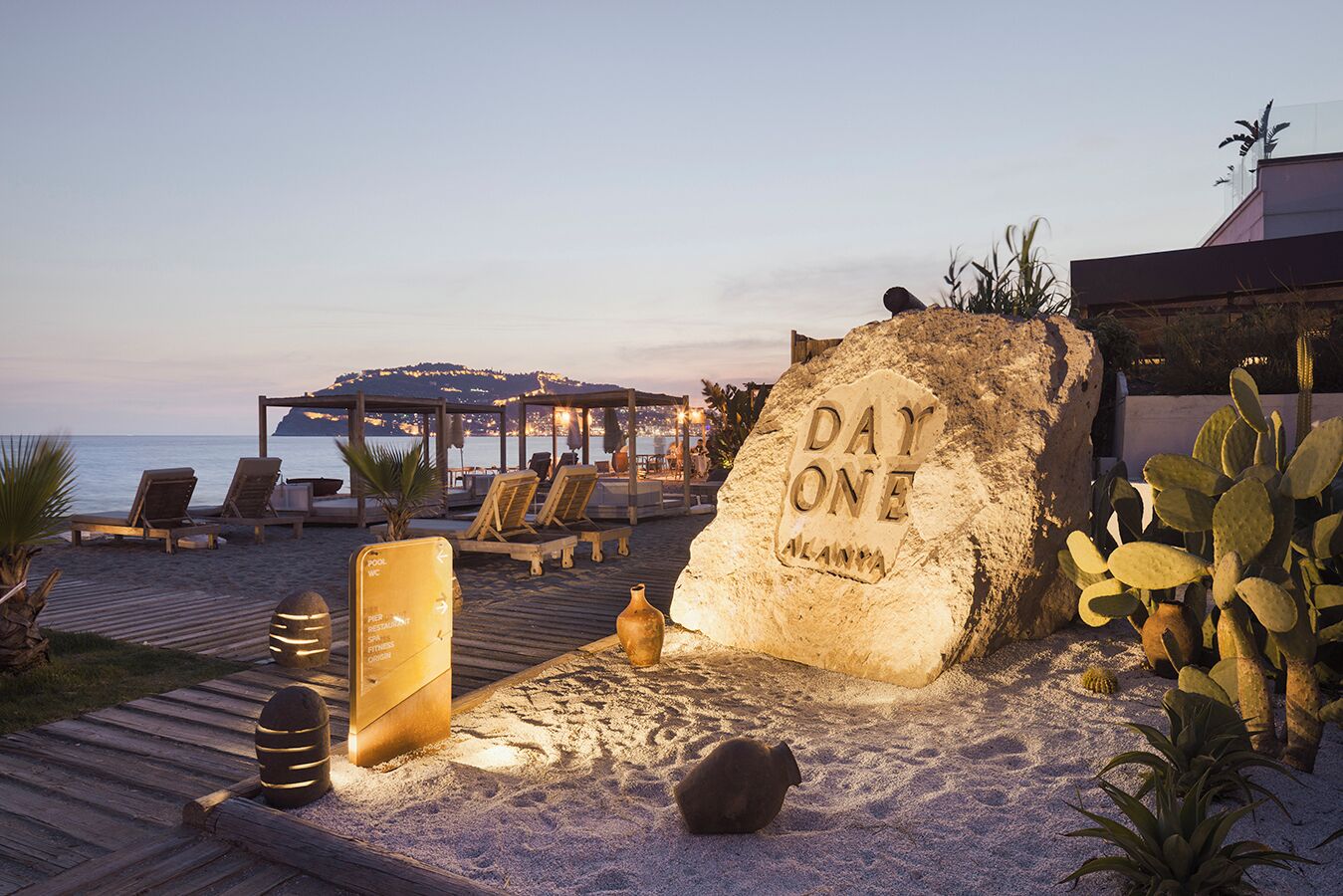 A beachside lounge area with sun loungers, a wooden deck, and a large rock with "DAY ONE ALANNA" engraved on it.