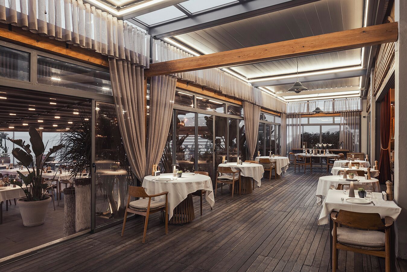Een restaurant met houten vloeren, tafels met witte tafelkleden en grote ramen met gordijnen.