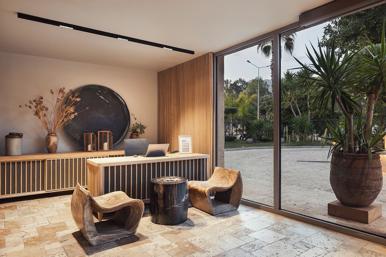 A modern reception area with a wooden reception desk, two lounge chairs, a large potted plant, and a view of the outdoor garden.