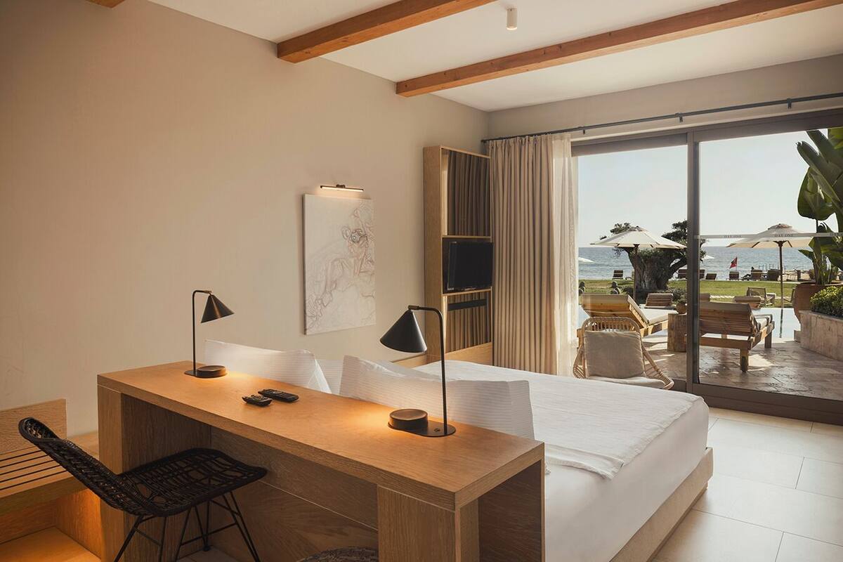 Une chambre d’hôtel moderne avec un bureau en bois, deux lampes noires, un lit et une grande porte-fenêtre coulissante donnant sur un balcon équipé de mobilier d’extérieur et offrant une vue sur la mer.