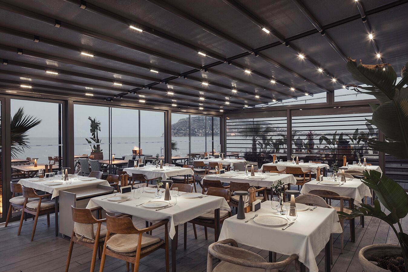 Ein Restaurant mit Außenbereich, Tischen zum Essen, großen Fenstern mit Meerblick und einem modernen Deckenstil.