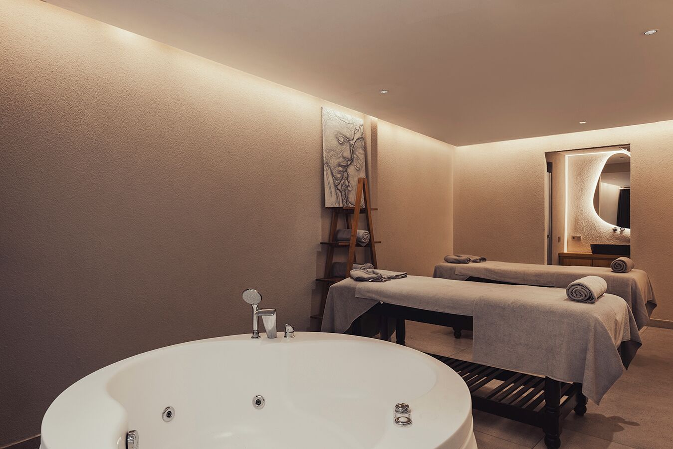A modern bathroom with a large bathtub, two massage tables, and a wooden shelf with towels.