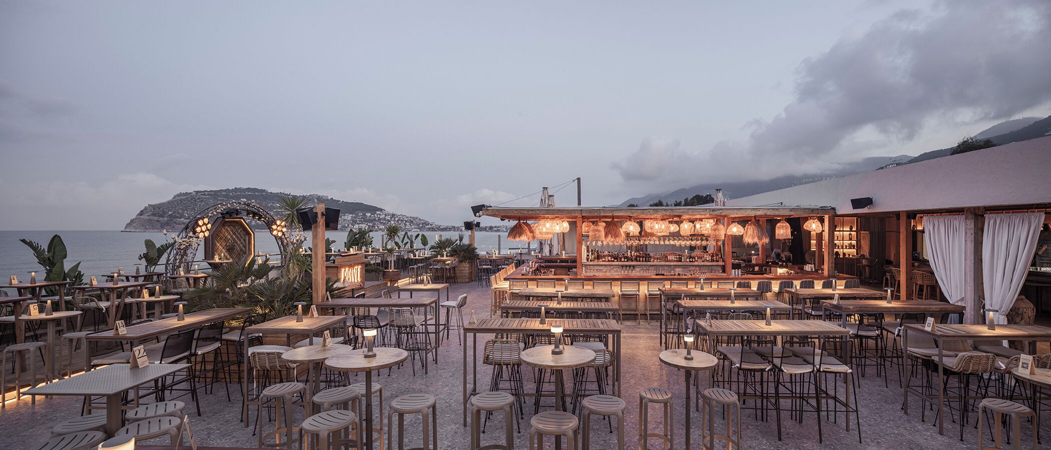 Un restaurante al aire libre con mesas, sillas, una zona de bar y vistas al mar y a la montaña.