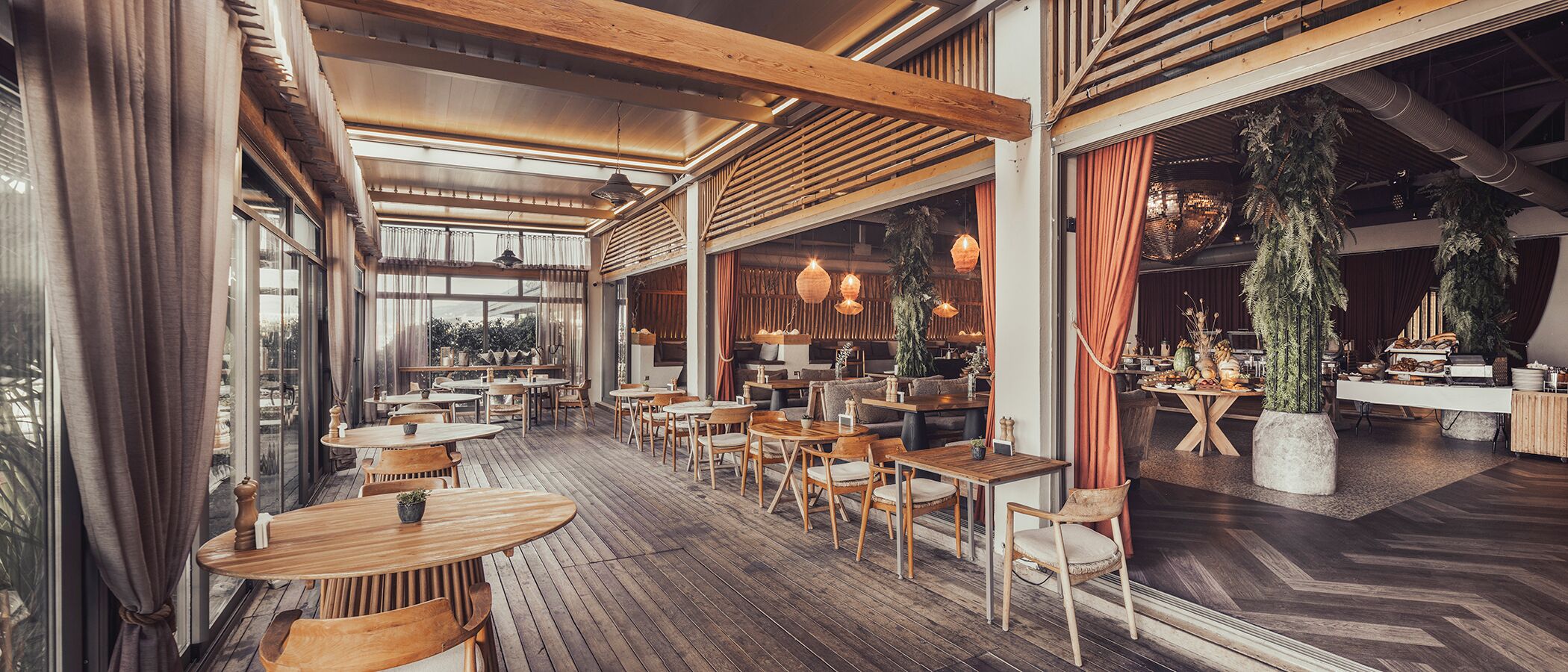 Un restaurant avec des tables et des chaises en bois, de grandes fenêtres et des plantes suspendues.