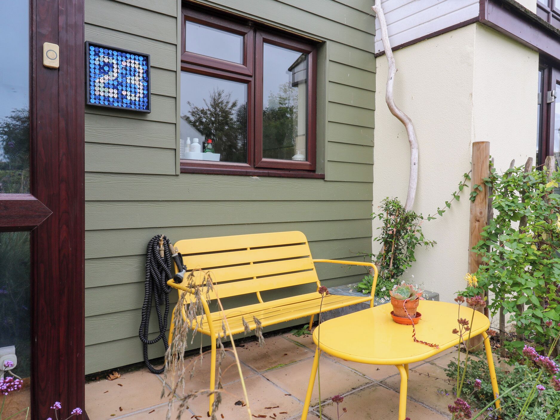 A yellow bench and table outside a house with a window and a blue mosaic on the wall.