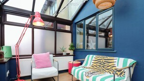 A cozy room with a blue wall, a window, a chair with a pink pillow, a striped sofa, a red floor lamp, and a green watering can.