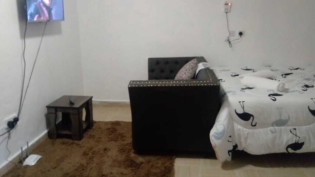 A bedroom with a bed, a small table, a television mounted on the wall, and a power outlet. - Stay.Plus Utawala Studio Apartment (Nairobi)