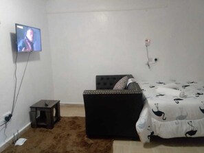 Un dormitorio con cama, una mesita, televisor montado en la pared y toma de corriente.