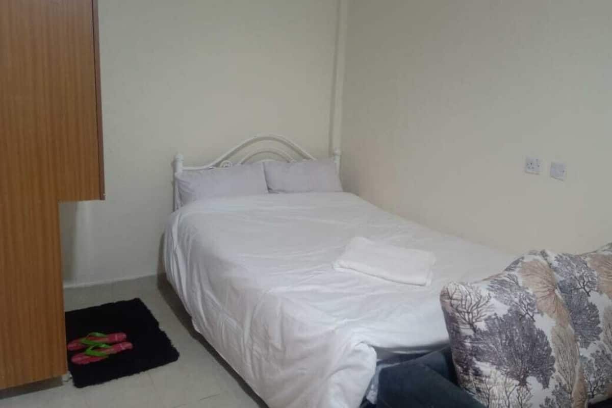 A single bed with white bedding, a black rug with pink slippers, and a patterned throw on the right.