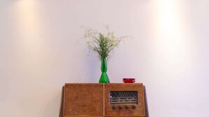 Ein altes Radio mit einer grünen Vase und einem roten Gegenstand darauf, vor einer schlichten Wand mit zwei Wandleuchten.
