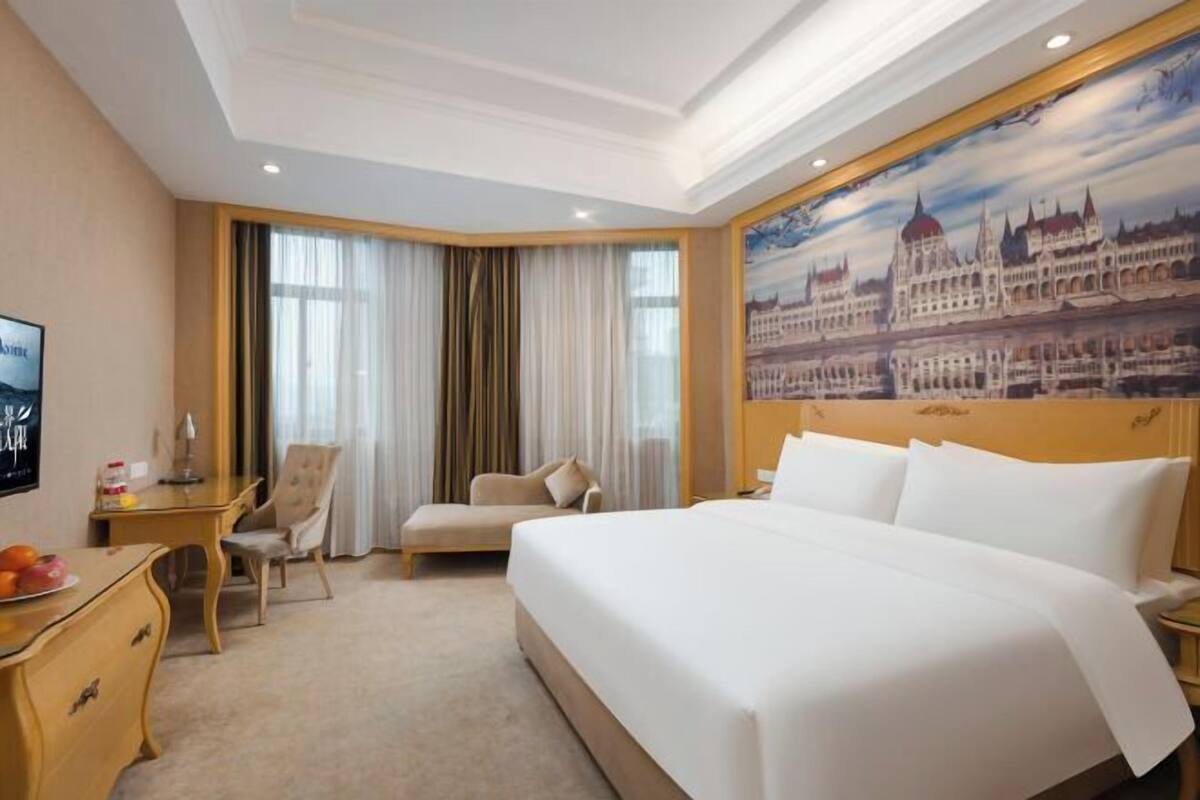 A hotel room with a large bed, a desk, a chair, and a painting of a building on the wall.