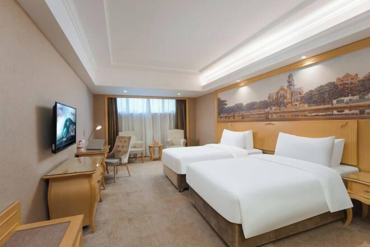 A hotel room with two beds, a desk, a chair, a TV, and a painting on the wall.