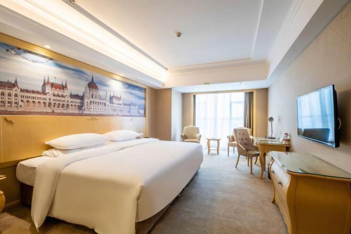 A hotel room with a large bed, a desk, a chair, a television, and a painting of a building on the wall.