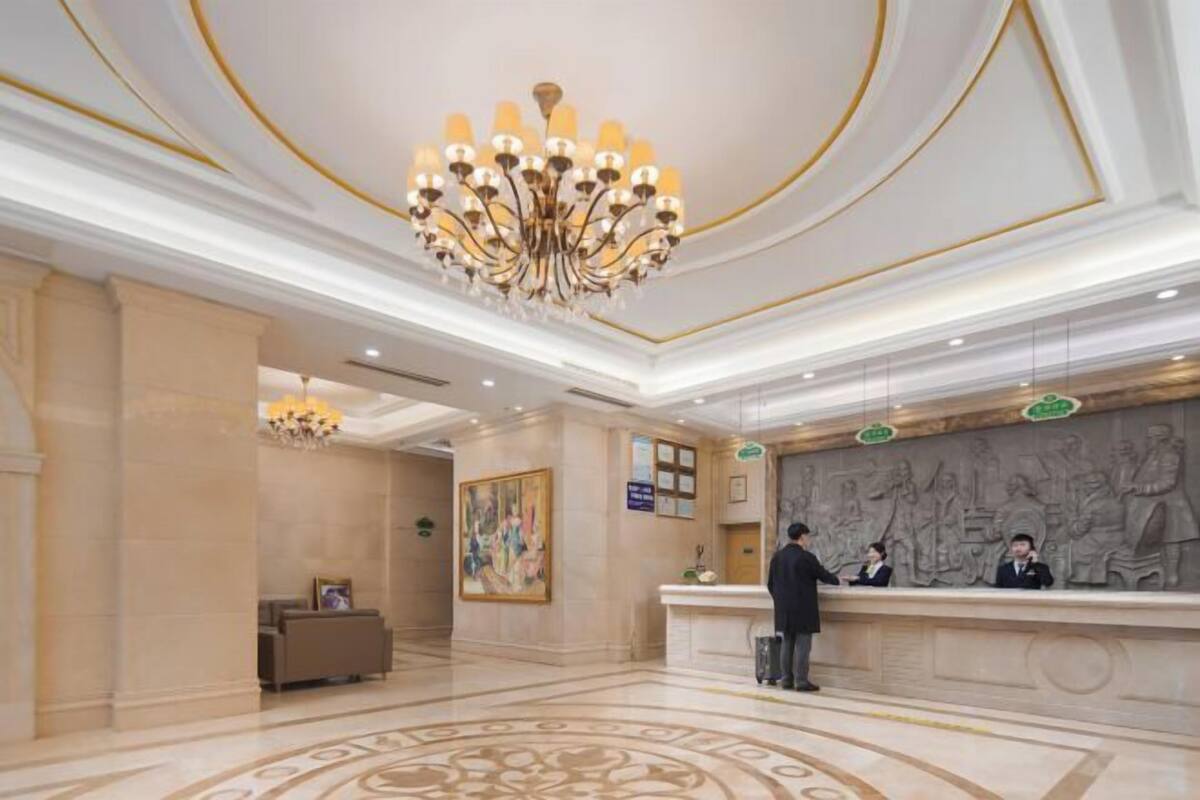 A hotel reception area with a marble floor, a large chandelier, a receptionist desk, and a wall with a relief sculpture.