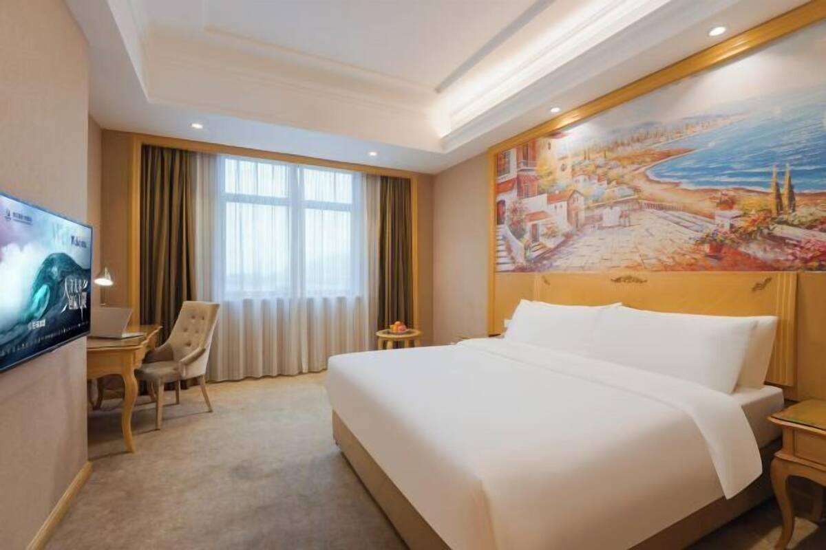 A hotel room with a large bed, a desk with a chair, a television, and a painting on the wall.
