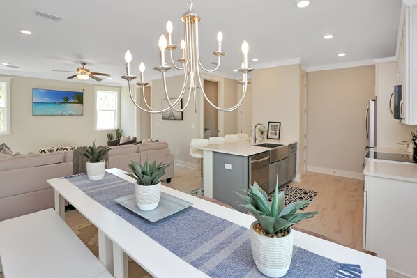 A modern kitchen with a central island, a dining area with a long table, and a living room with a sofa and a ceiling fan.