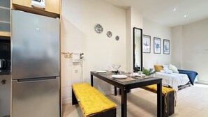 Una cocina compacta con refrigerador, una mesa de comedor con bancos y una cama al fondo.