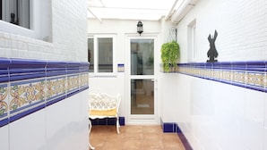 Un estrecho pasillo con paredes de azulejos blancos y bordes decorativos azules, un banco blanco, y una puerta que da a una habitación.