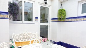 Una mesa blanca con detalles ornamentales, una silla blanca, una planta colgante y una ventana con detalles azules.