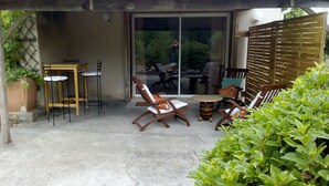 An outdoor patio area with wooden furniture, including a table and chairs, and a glass door leading to a garden. - Charming Gîte (4 people) ideal for couples or families, near Montcuq
 (PORTE DU QUERCY)