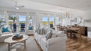 A spacious living area with a white sofa set, a wooden dining table, and a ceiling fan. Large windows provide a view of the outdoors.